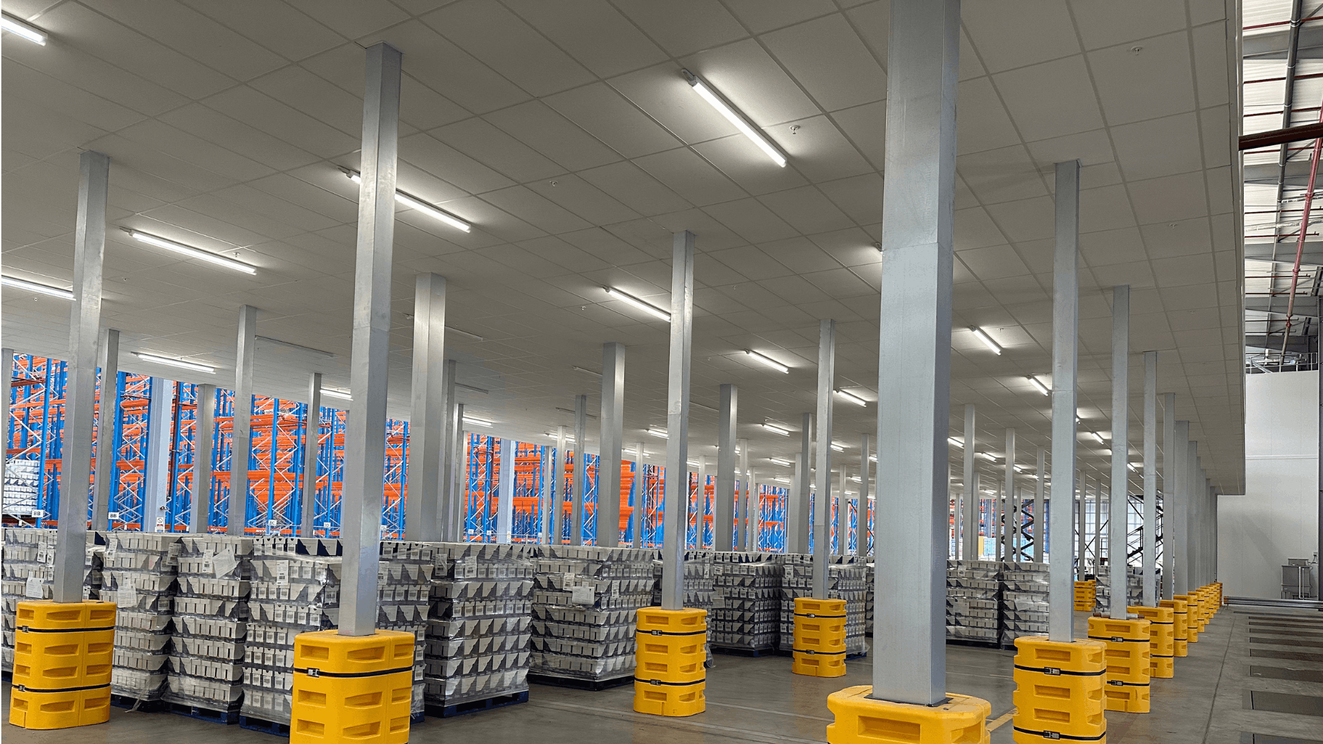 Ground floor storage area beneath Ziegler mezzanine floor, showing fire-rated ceiling, lighting, steel columns, palletised goods and yellow column protectors